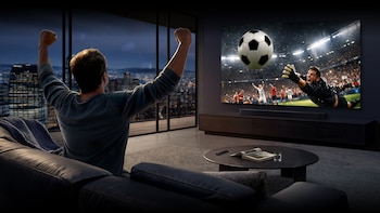 Un hombre de espaldas, con los brazos en alto, mira un partido de fútbol en un televisor Samsung grande en un apartamento con vistas a una ciudad nocturna