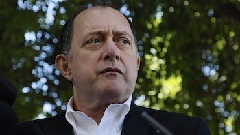 Hombre de mediana edad con cabello oscuro y traje oscuro sobre camisa blanca mira hacia la derecha, con un fondo verde borroso de hojas de árboles