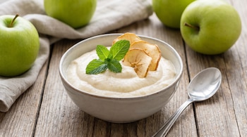 Un bol de mousse de manzana, hojas de menta, rodajas de manzana deshidratada, cuchara de metal, varias manzanas verdes y un paño claro sobre una mesa de madera.