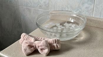 Un bol transparente con agua y cubitos de hielo. Una diadema de tela rosa con un lazo. Una encimera clara y azulejos en la pared.