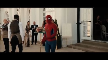 Un hombre disfrazado de Spider-Man, varias personas incluyendo camareros, una mesa con arreglo floral, un edificio blanco y una puerta abierta