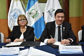 Una mujer y un hombre sentados frente a una mesa firman documentos con las banderas de Guatemala a sus espaldas