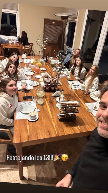 Hombre sonriente en primer plano. Un grupo de niñas y una mujer sentadas alrededor de una mesa de madera con platos, vasos, jarras y postres. Texto: "Festejando los 13!!!"