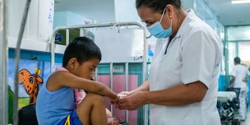 Enfermera con mascarilla azul y uniforme blanco asiste a un niño con camiseta azul sentado en una cama de hospital con barandilla y mural infantil