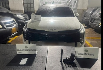 Un vehículo blanco estacionado detrás de una mesa que contiene un teléfono celular, una pistola con cargador, munición y letreros