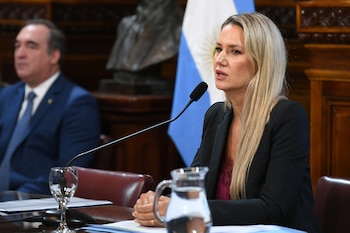 Carolina Losada, con cabello rubio y chaqueta oscura, habla en un micrófono con la bandera argentina de fondo y un hombre en segundo plano