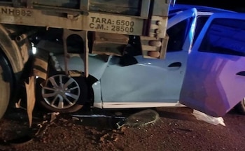Primer plano de un coche azul claro dañado y parcialmente bajo un camión de carga por la noche, con la puerta abierta y fragmentos de vidrio en el suelo