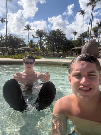 Dos hombres jóvenes en una piscina. Uno sonríe a cámara, el otro usa gafas de sol. Palmeras y cielo azul con nubes en el fondo