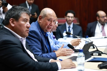 Vista de cerca de varios hombres sentados en una mesa de reuniones con micrófonos y botellas de agua, uno de ellos hablando y gesticulando