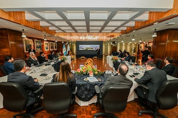 Sala de conferencias con mesa en forma de U, personas sentadas, un orador frente a una pantalla con vista aérea urbana, paredes de madera y cuadros