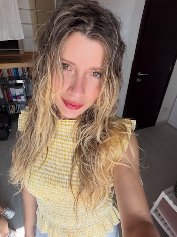 Una mujer con cabello rubio ondulado y top a cuadros amarillos mira hacia la cámara. Detrás, se ven un estante con libros y una puerta oscura