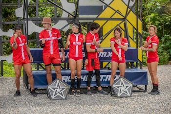 Seis atletas del equipo rojo de Exatlón México, con uniformes rojos y blancos, posan en un campo de grava con estructuras geométricas y árboles de fondo