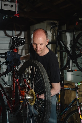 Un hombre repara una bicicleta roja con neumáticos negros y rayos plateados. Se observan otras bicicletas al fondo y un motor de garaje