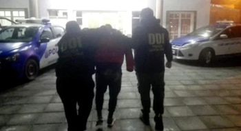 Tres hombres, dos de ellos con chalecos de la DDI, escoltan a un tercero frente a un edificio con dos patrulleros policiales a los costados