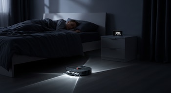 Una aspiradora robot negra ilumina el suelo oscuro de un dormitorio. Una persona duerme en la cama adyacente y un reloj digital marca las 3:00 AM.