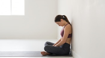 Mujer con top deportivo y leggings sentada en una colchoneta contra una pared blanca, con la cabeza inclinada hacia el pecho.