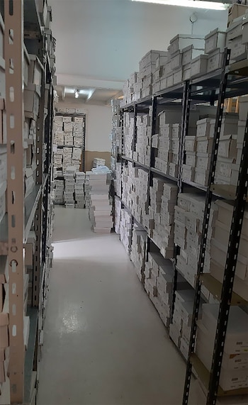 Pasillo estrecho con estanterías metálicas a ambos lados y al fondo, llenas de cajas rectangulares blancas apiladas