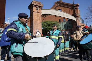 Un grupo de hombres, algunos con uniformes azules y turquesas, camina con tambores grandes frente a un arco de ladrillos que dice Parque de la Estación