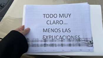 Una mano sostiene un cartel blanco con el texto impreso en negro 'TODO MUY CLARO... MENOS LAS EXPLICACIONES', con manchas de tinta en la parte inferior