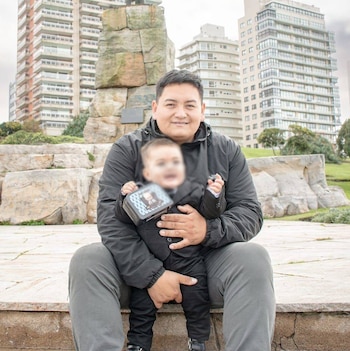 Hombre sonriente sentado con un niño en brazos en un parque de la ciudad. Edificios altos y un monumento rocoso se ven al fondo