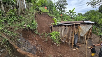 Vista de un deslizamiento de tierra cerca de una casa precaria de madera y lámina, rodeada de vegetación tropical exuberante en una ladera, con figuras humanas en la distancia