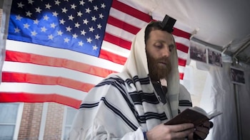 Un hombre con barba, tallit blanco y negro y tefilín negro en la cabeza, sostiene un libro mientras la bandera de Estados Unidos se ve detrás
