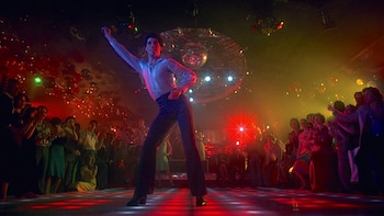 Un hombre con camisa blanca y pantalones oscuros baila sobre una pista iluminada con luces rojas, verdes y amarillas, rodeado de gente y una bola de discoteca en el fondo