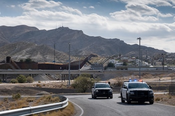 Dos camionetas policiales circulan por una carretera de asfalto frente a un muro metálico y montañas al fondo.