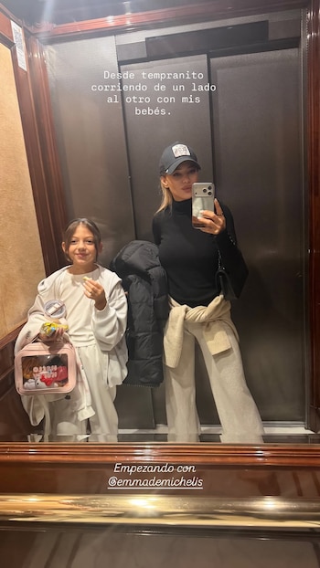 Una mujer toma una foto frente al espejo de un ascensor junto a una niña que sostiene una bolsa rosa