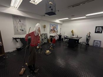 Una sala de exposición con piso negro y paredes blancas muestra varias esculturas figurativas hechas con ropa y bolsas plásticas, y obras de arte en las paredes