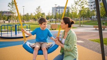 Una mujer de camisa verde y un niño de camiseta azul y pantalones cortos están en un columpio de un parque infantil con suelo amarillo y azul.