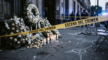 Cinta amarilla con la leyenda escena del crimen frente a coronas fúnebres blancas, velas encendidas, marcas de tiza y sillas metálicas.