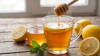Una taza de té caliente con rodajas de limón y vapor, un tarro de miel con un cucharón vertiendo, limones y menta en una mesa de madera.