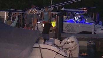 Personas en varias embarcaciones amarradas a un muelle en la noche. Se observa un motor fueraborda y luces azules en un barco