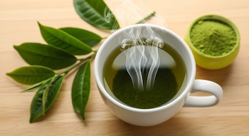 Una taza blanca con té verde caliente y vapor, acompañada de hojas de té verde frescas y un tazón con polvo de matcha, sobre una superficie de madera clara.