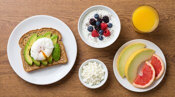 Tostada con aguacate y huevo poché en plato blanco, tazón de yogur con frutos rojos y semillas de chía, jugo de naranja, queso cottage, rebanadas de melón y pomelo.