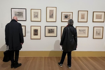Vista trasera de dos personas, una calva con abrigo oscuro y otra con cabello gris y chaqueta de cuero, mirando grabados enmarcados en una galería de arte