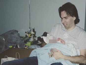 Steven Pladl, un hombre de cabello oscuro, sostiene a un bebé envuelto en una manta azul. Se ven un peluche y un arreglo floral al fondo