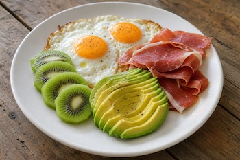 Plato blanco con dos huevos fritos, lonchas de jamón serrano, medio aguacate rebanado y rodajas de kiwi sobre una mesa de madera.