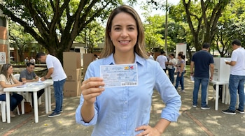 Mujer sonriente de cabello castaño con camisa azul claro, posando al aire libre y mostrando un certificado electoral. Al fondo, se observan personas y árboles.