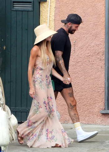 Mujer con vestido floral largo, sombrero de paja y pelo rubio camina de la mano con hombre con gorra negra, camiseta negra, shorts y tatuajes visibles