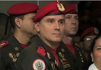 Oficial militar con uniforme oscuro, boina roja y medallas, acompañado de otros soldados de uniforme y boina, y una mujer con parte del rostro visible