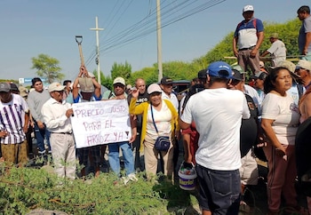 Manifestantes, en su mayoría agricultores, bloquean una carretera con ramas y exhiben un cartel blanco que dice "PRECIO JUSTO PARA EL ARROZ YA!". Un hombre sostiene una pala en alto