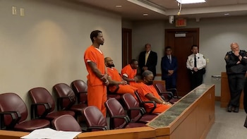 Grupo de hombres con uniformes naranjas y esposas, algunos de pie y otros sentados en bancos de sala de justicia, con oficiales y personas vestidas de traje al fondo