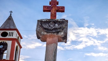 Cruz de piedra rojiza con relieves de figura religiosa y ángel, sobre pedestal. Campanario de iglesia blanco y rojo con cruz. Cielo azul con nubes