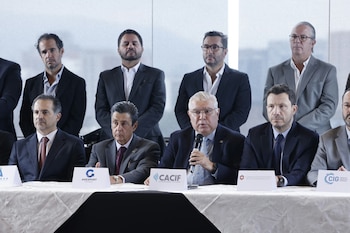 Hombres en trajes sentados en una mesa con micrófonos y letreros de organizaciones como CACIF y AGEXPORT. Otros hombres observan de pie detrás