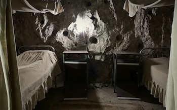 Habitación excavada en roca con paredes irregulares, dos camas individuales con sábanas claras, dos mesitas de noche oscuras y una luz central en la pared