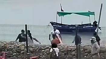 Varias personas pelean cerca de una reja en una playa con piedras mientras una embarcación con toldo verde permanece en el agua