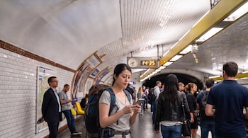 Una mujer con mochila sostiene un teléfono inteligente en el andén de una estación de metro subterránea con otros pasajeros al fondo.