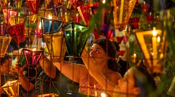 Una mujer coloca un farol azul encendido en una estructura metálica rodeada de faroles multicolor con velas en su interior.